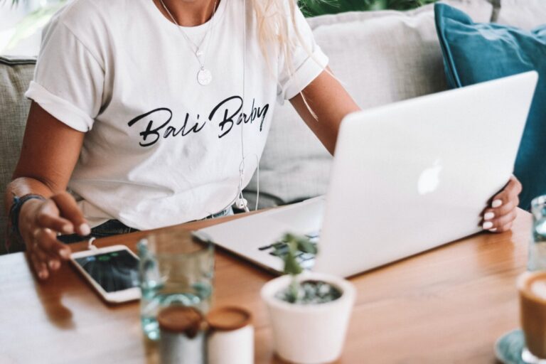A person wearing a Bali Baby T-shirt is sitting at a wooden table, using a laptop. A smartphone, glasses of water, small jars, and a potted plant are also on the table. Earbuds are visible, and the setting appears relaxed and casual.