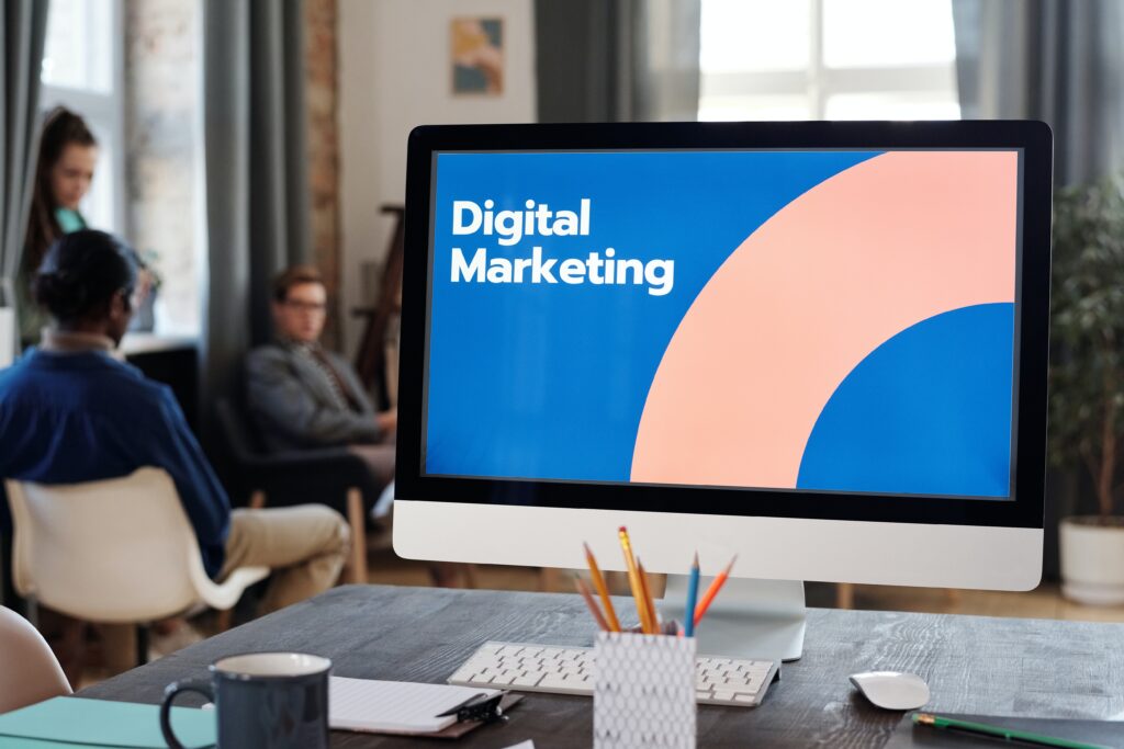 A computer monitor displays the text Digital Marketing with graphic elements. In the background, a group of people are sitting in a room with large windows, engaging in a discussion. A desk with pencils and a notebook is in the foreground.
