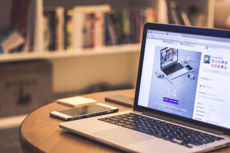 A sleek laptop displaying a webpage featuring a WordPress theme is placed on a wooden table in a cozy room. Nearby, a smartphone and a pen complete the setup. Books are neatly arranged on a bookshelf in the blurred background.