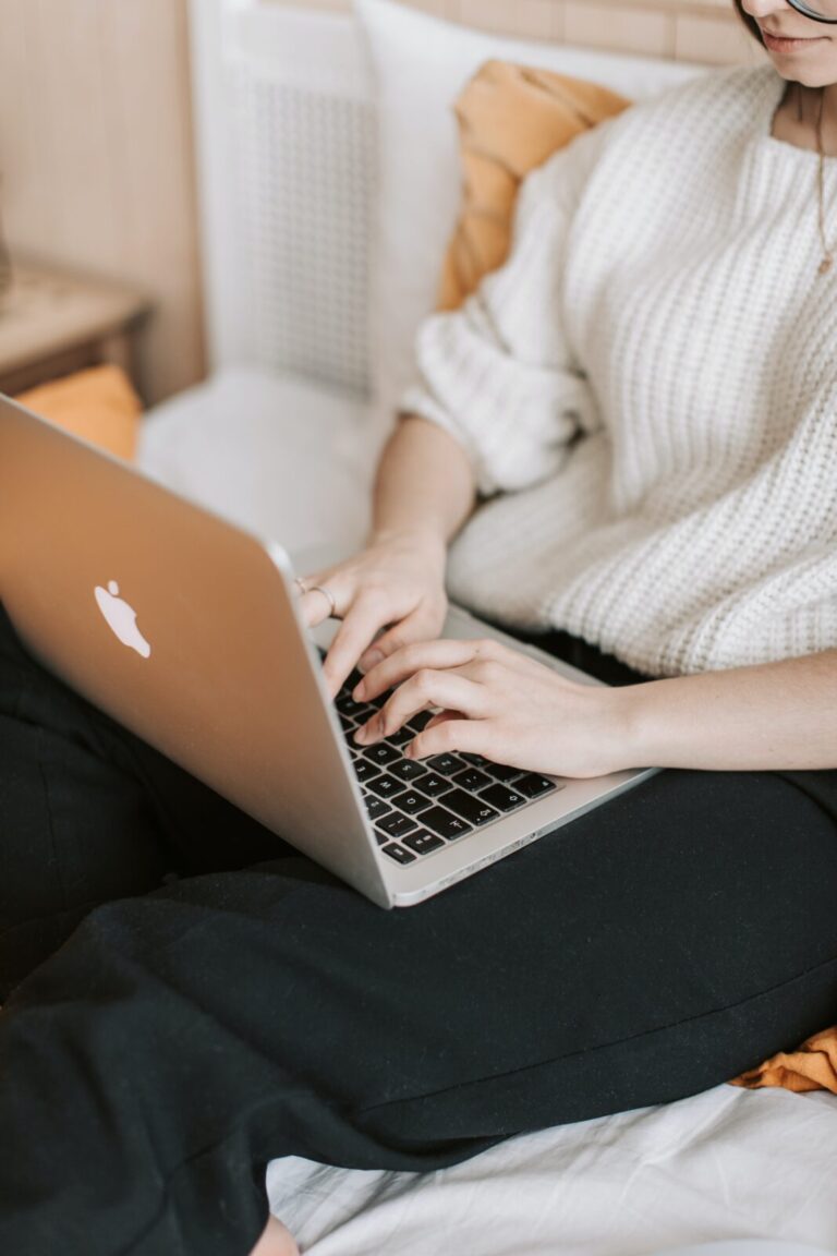 Person wearing a white sweater and black pants is sitting on a bed, typing on a silver laptop. The room has a cozy ambiance with soft lighting and earth-tone bedding.