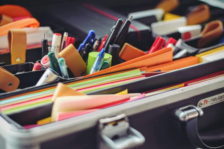 An open briefcase filled with assorted office supplies, including colorful files, pens, and markers. The items are neatly arranged, creating a vibrant array of colors against the metallic finish of the briefcase.
