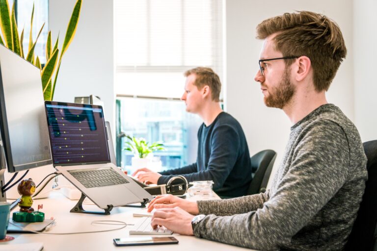 Two men work at desks with laptops in a bright office. One man in the foreground focuses on a computer screen, while the other sits behind him. Plants and office supplies are visible. Light streams in through a window.