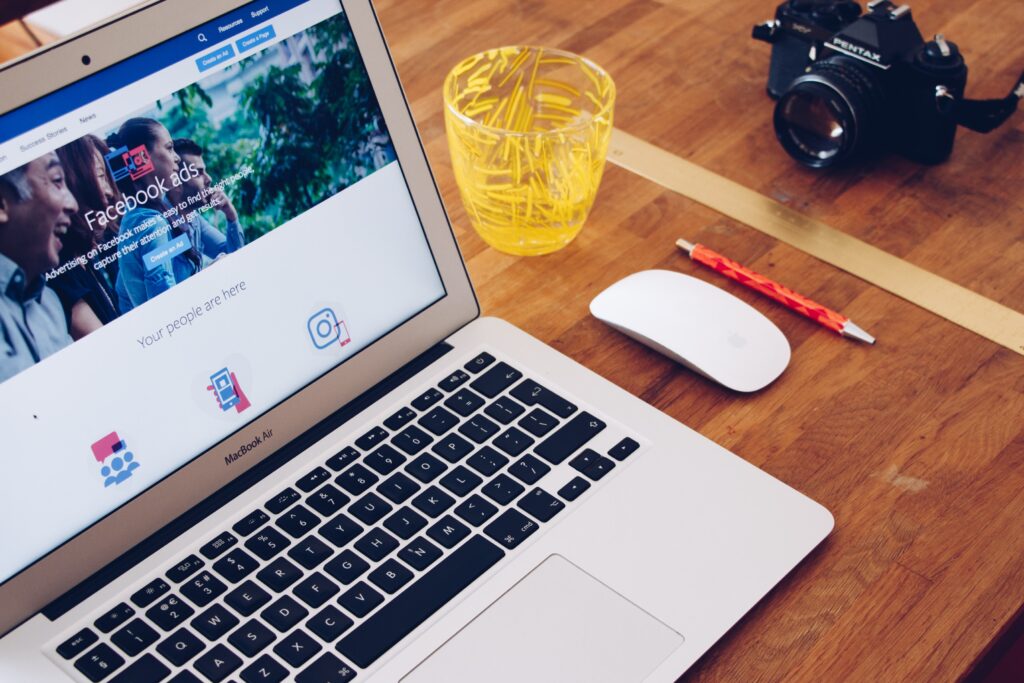 A laptop displaying a Facebook ads page is on a wooden table. Nearby, there is a wireless mouse, a pencil, a yellow transparent cup with paper clips, and a camera.