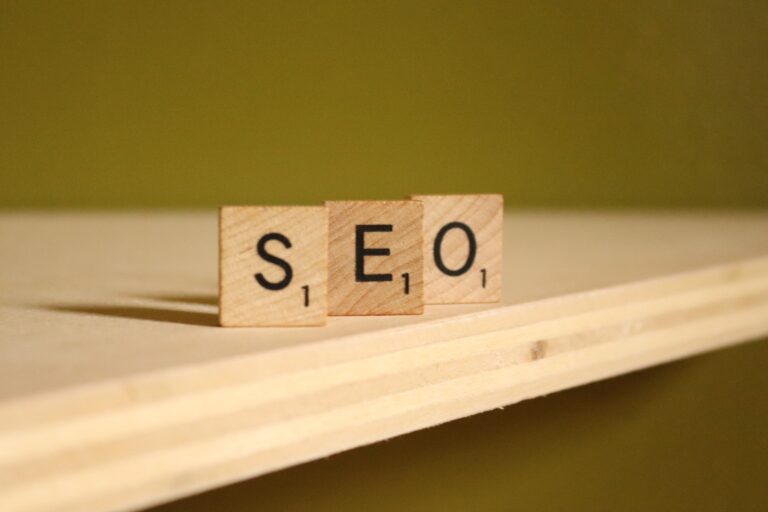 Wooden letter tiles spelling SEO are arranged in a row on a light wooden surface with an olive green background. Each tile shows the letter and the number 1 in a Scrabble-like style.