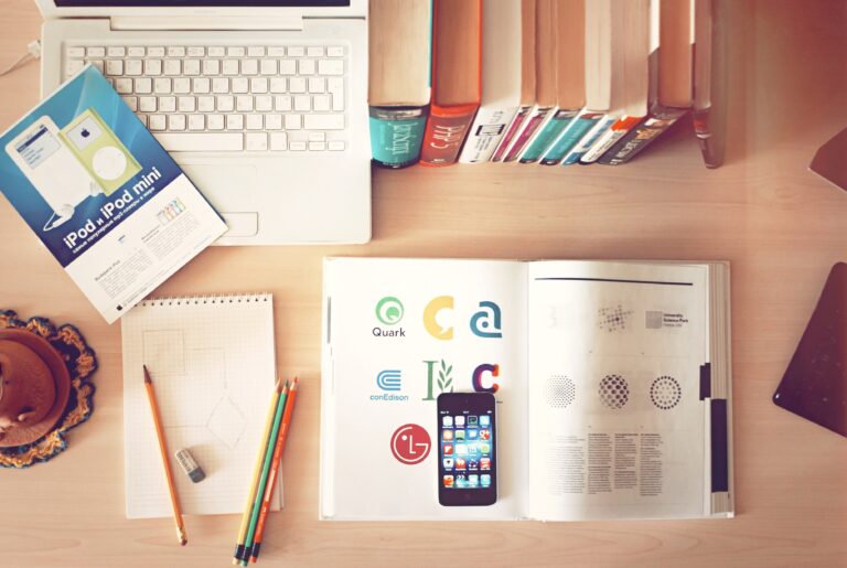 A neatly organized desk with an open laptop, a book with colorful logos, a smartphone, a stack of books, and some stationery items like pencils and a notepad, all on a light wooden surface.
