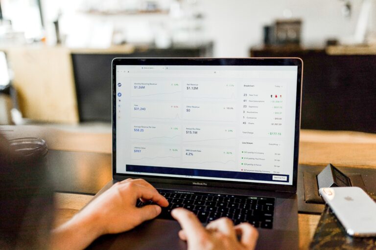 A person using a laptop displaying analytics and graphs on the screen. It shows data metrics such as revenue, conversions, and performance indicators. The laptop is placed on a wooden table in a well-lit room with blurred background elements.