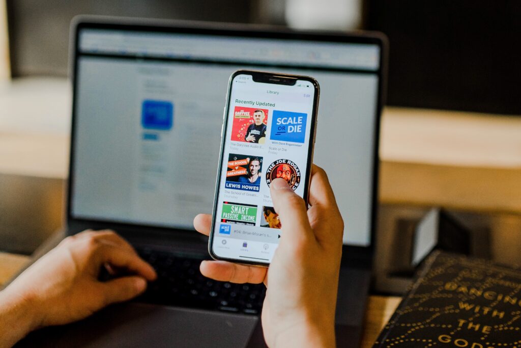 A person uses a smartphone while seated at a laptop. The phone screen shows various podcast covers. The laptop screen is blurred. There is a book on the desk next to the laptop.