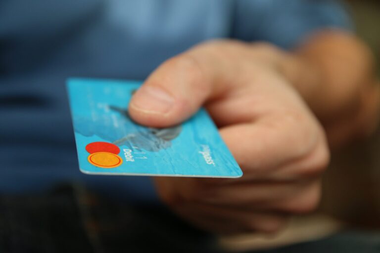 Close-up of a hand holding a blue debit card with visible logos. The background is blurred, focusing on the card and hand.