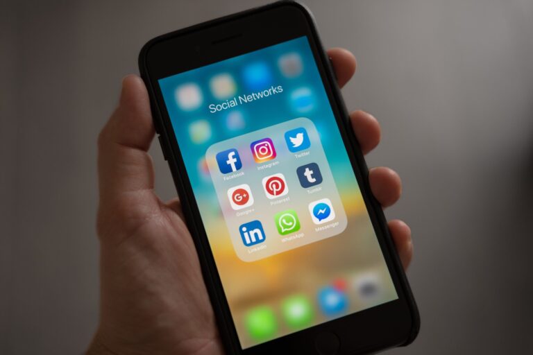 A hand holds a smartphone displaying a folder titled Social Networks on the screen. Inside the folder are app icons for Facebook, Instagram, Twitter, Google+, Pinterest, Tumblr, LinkedIn, WhatsApp, and Messenger.