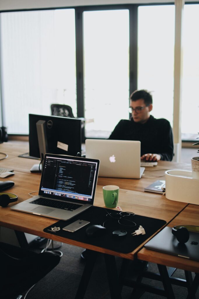 An open office space with two people working on laptops at wooden desks. One laptop displays code. The desks are equipped with various office supplies, and there is a green mug near the laptops. Large windows provide natural light.