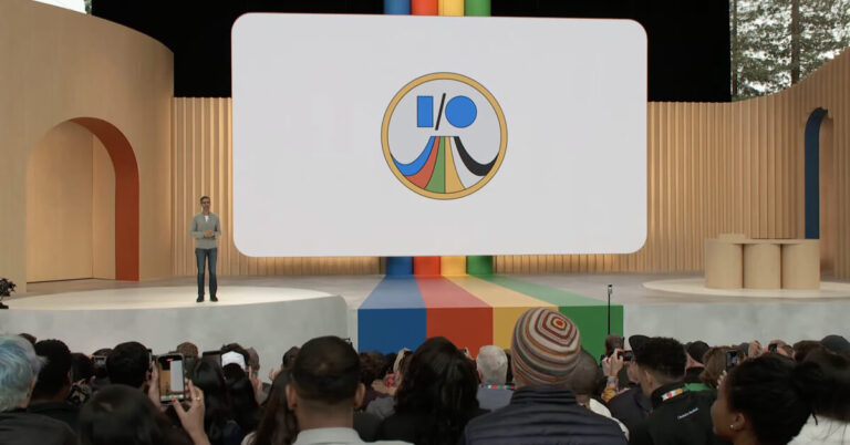 A person stands on a stage with a large screen displaying a circular logo featuring stylized geometric shapes. The audience, some holding phones, closely watches the presentation. The stage has colorful, curved design elements.